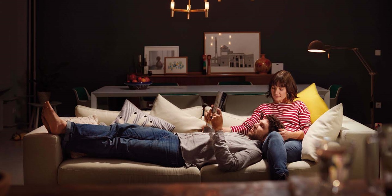 Person relaxing on a sofa with another person using a tablet in a living room with modern lighting fixtures